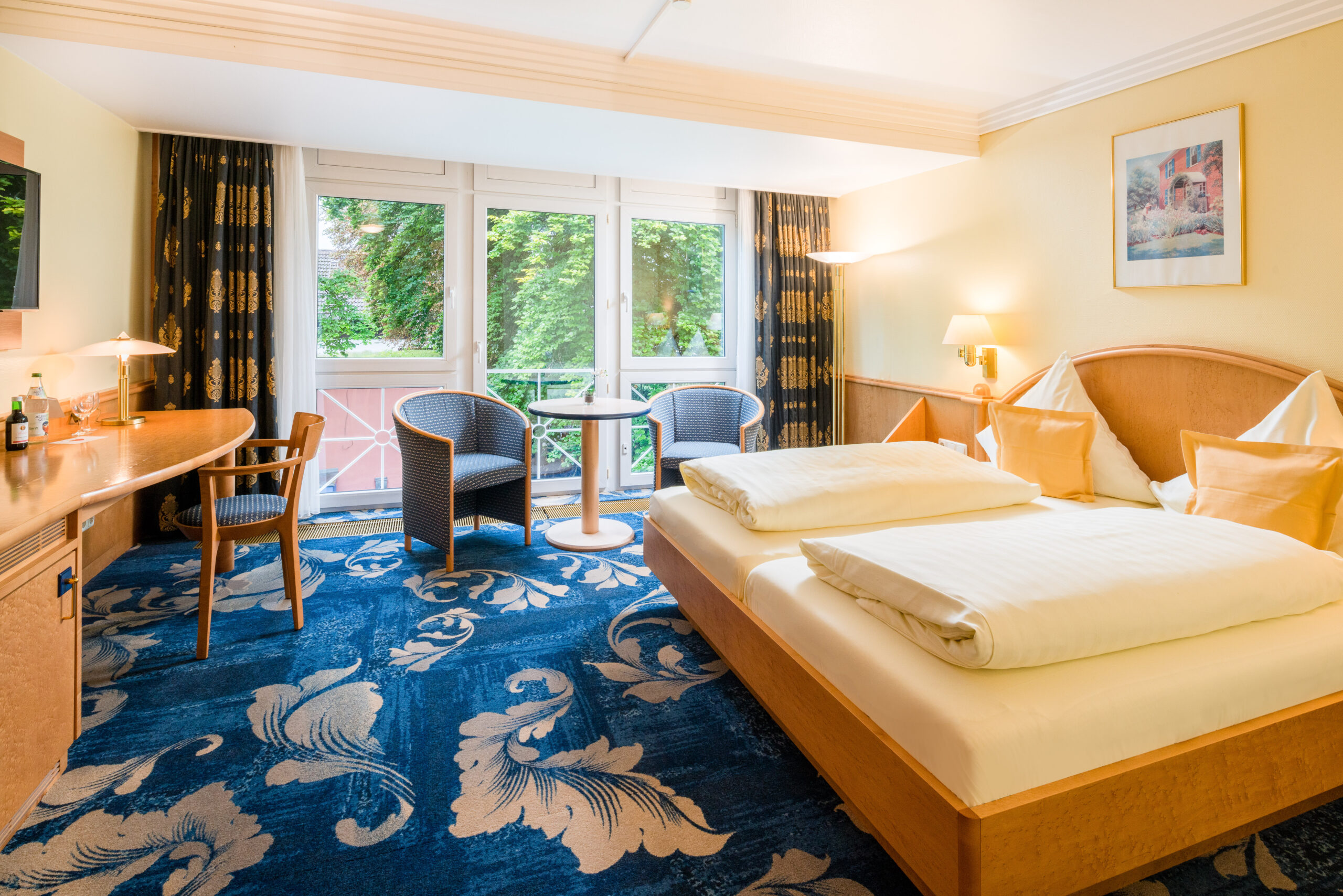 Hotel Asemann Planegg Comfort Double Room with Garden View A light-filled Comfort double room at the Hotel Asemann in Planegg, featuring a large double bed, a cosy seating area, a patterned blue carpet and floor-to-ceiling windows overlooking the countryside.