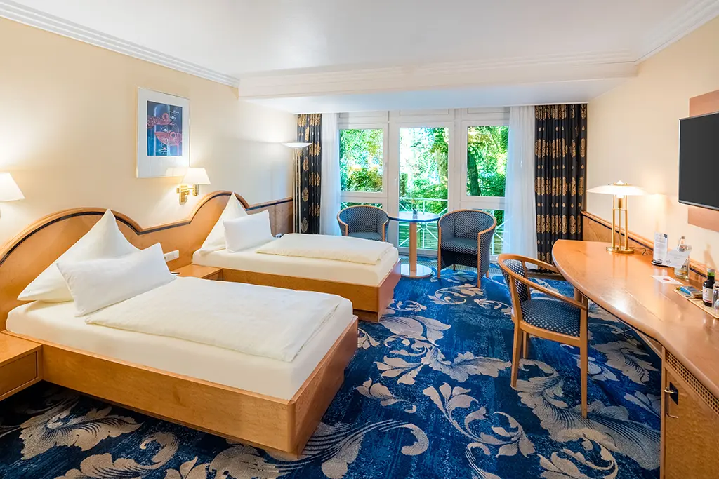 Hotel Asemann Planegg Comfort Double Room with Garden View Spacious Comfort double room at the Hotel Asemann in Planegg, featuring a large double bed, a blue patterned carpet, a seating area and floor-to-ceiling windows overlooking the green garden.